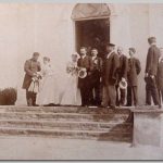 Bruidspaar met meerdere mannen (sommigen in uniform) en een dame voor de ingang van een kerk