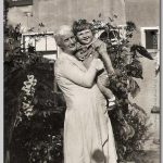 Dame met een klein kind op de arm achter een huis in de tuin.