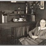 Lydia, 1927 Jonge vrouw in een stoel bij een meubel met kopjes en boeken er op