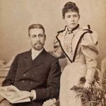 Portret in een studio van een echtpaar. De man zit inheeft een krant in de handen. De vrouw staat naast hem