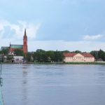 breed water met aan de overkant een kerk en huizen. Links een gedeelte van de brug